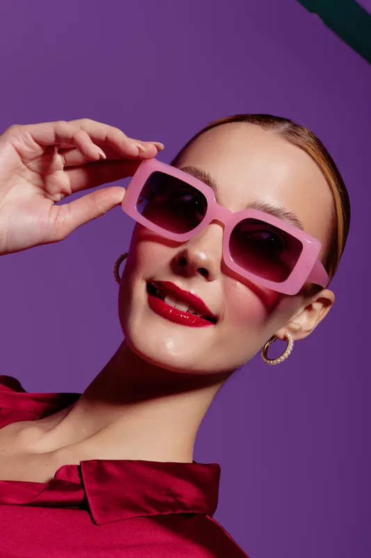 Young woman with pink sunglasses and red lipstick poses against purple background. Fashion portrait with stylish accessories and confident expression. Concept of beauty and style.