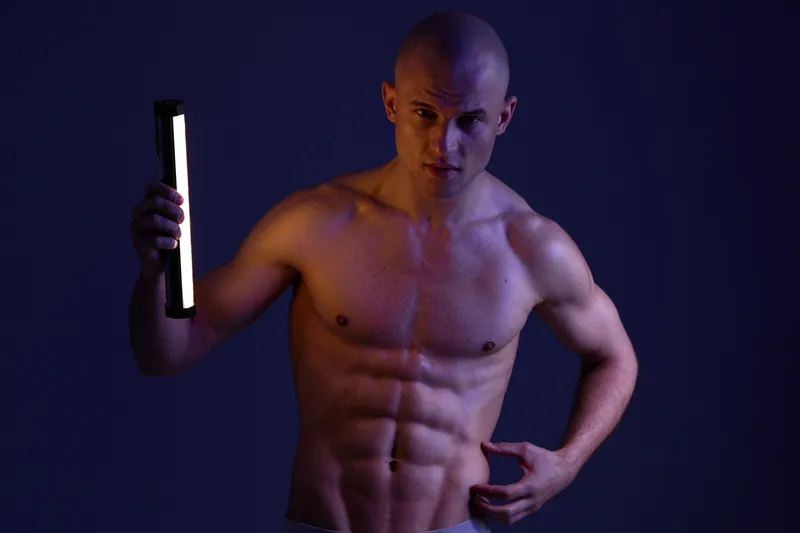 Portrait of a muscular man holding a glowing light stick, highlighting his toned physique against a moody backdrop. Evokes strength and determination in dark lighting.