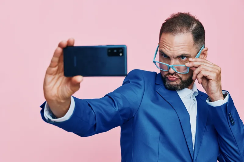 A focused business professional takes a bold selfie against a vivid pink background, revealing confidence and curiosity as he adjusts glasses and captures a dynamic moment.