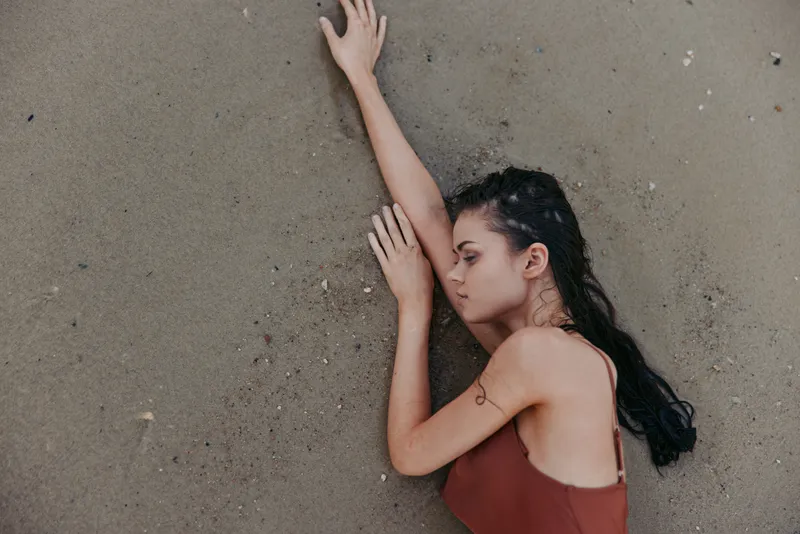 A lone woman lies on sandy beach, eyes closed, arms stretched along her body, wearing a rust-colored dress, conveying stillness, fatigue, and contemplative mood.