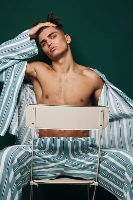 Young man wearing open striped pajama set sitting behind metal chair shirtless while posing with hand on his head against green background in interior studio lighting.