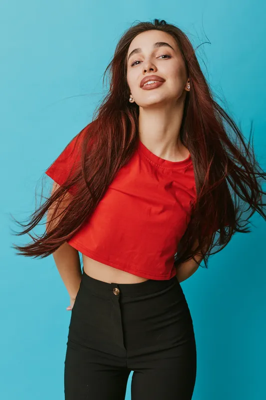 Young woman with long hair wearing red crop top and black pants posing confidently against blue background. Casual style, fashion, beauty and happiness with smiling face and relaxed posture.