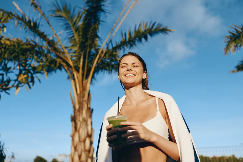 Young woman wearing a white bikini top with a towel over her shoulders enjoys a refreshing drink under a clear blue sky beside palm trees, her smile conveying a carefree mood.