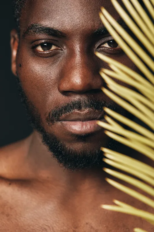 African man portrait with dark skin and beard in creative conceptual visual style holding palm leaf close to face with serious expression on black background.