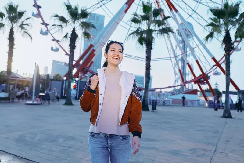 Happy woman strolling outdoors in an urban waterfront setting with palm trees and a ferris wheel, casual jacket and jeans, candid fashion, relaxed mood, sunny day, travel vibe, street scene