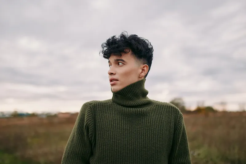 A candid portrait of a young man in an olive green turtleneck stands in an open field, conveying authenticity and quiet credibility under a muted cloudy sky.