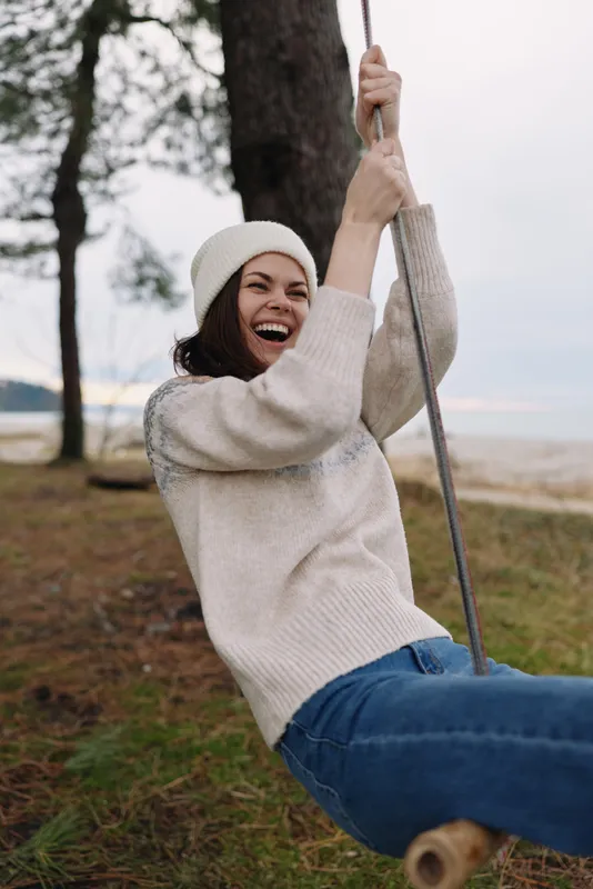 Woman on a swing outdoors, wearing a cozy sweater and beanie, smiling joyfully as she enjoys casual leisure in a sunny park setting