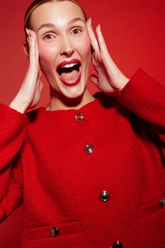 Excited woman in red coat with open mouth and hands on face expresses surprise and joy. Bright red background and warm lighting create energetic and festive mood for celebration concept.