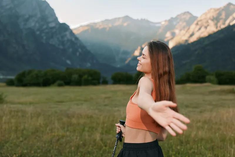 A joyful outdoor model is seen stretching her arms in a wide meadow with distant mountains. She wears an orange top and dark shorts, enjoying nature and a bright, carefree moment.