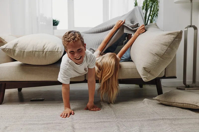 Happy children play and laugh under a blanket on a modern sofa in a bright living room, enjoying cozy family moments and playful interaction.