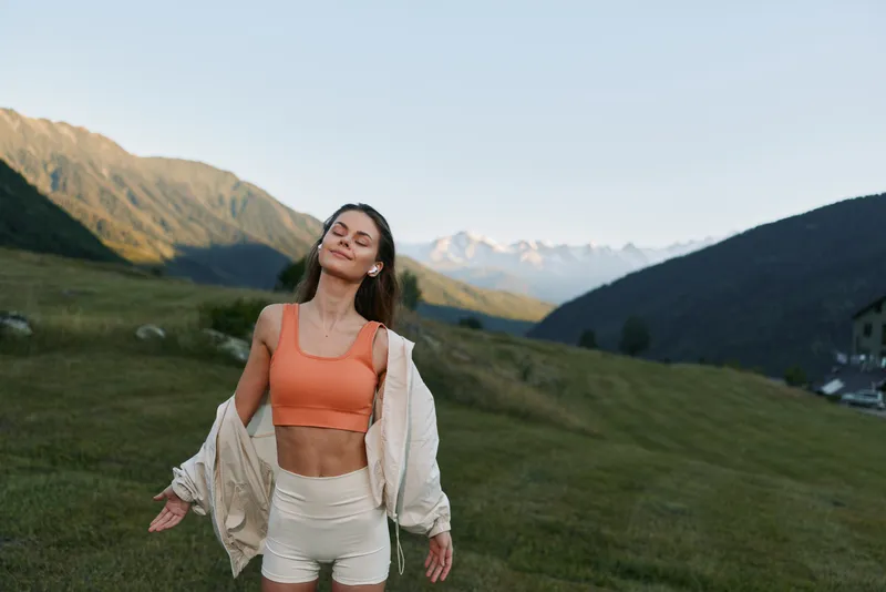 A young woman stands in a grassy valley with distant mountains, wearing an orange sports top and light jacket, enjoying fresh air, sunshine, and a carefree outdoor moment.