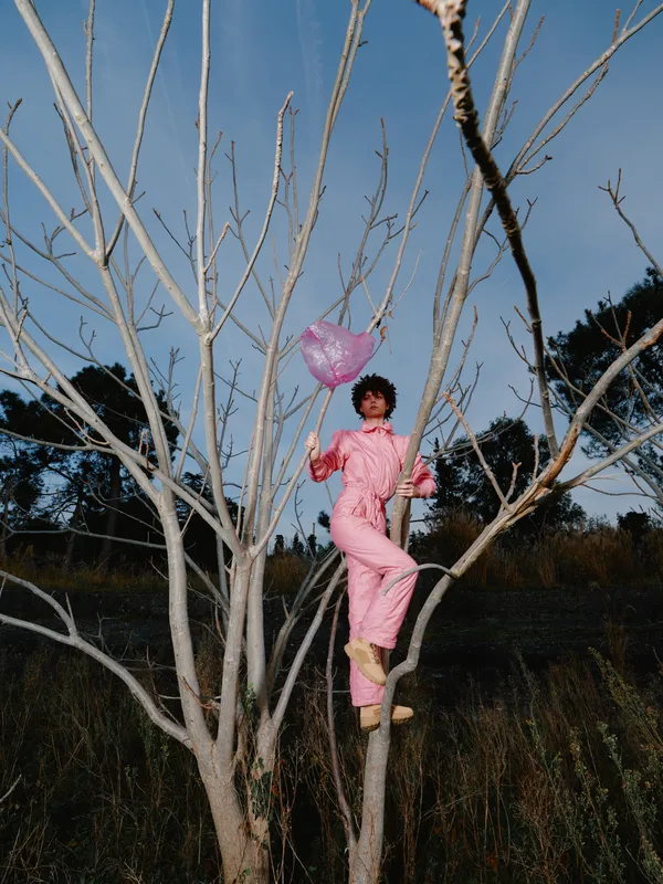 Woman climbing tree in pink jumpsuit holding a heart balloon in an outdoor scene nature setting, playful portrait among bare branches, capturing whimsical ascent and bold color