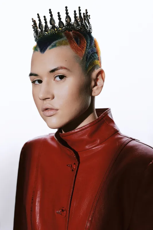 young woman with colorful hairstyle wearing red leather jacket and black decorative crown, posing confidently against plain white background, modern edgy fashion portrait