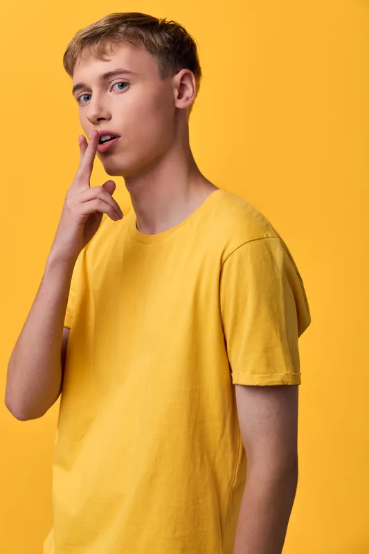 Thoughtful man in a yellow shirt stands against a matching yellow backdrop, finger to lips, conveying contemplation, curiosity, and focus in a vibrant, minimal studio setting.