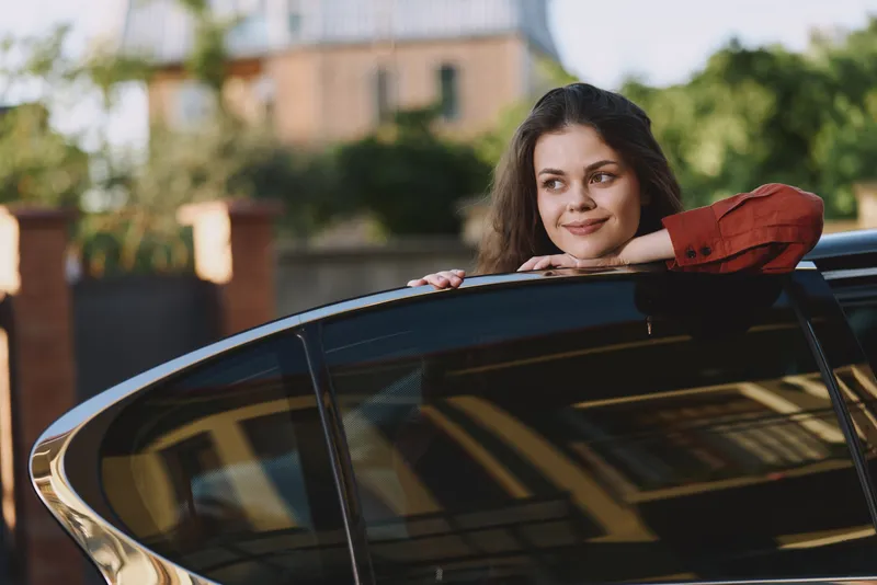 A young woman with dark hair leans on a glossy car window, smiling softly as sunlight highlights her relaxed pose and casual confidence