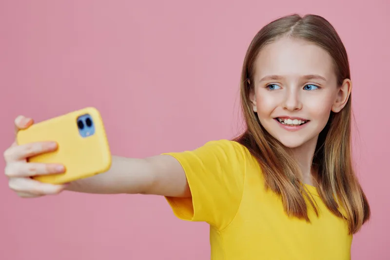 Happy young girl taking a selfie with a yellow phone against a pink background, showcasing joyful emotions and vibrant colors in a playful setting