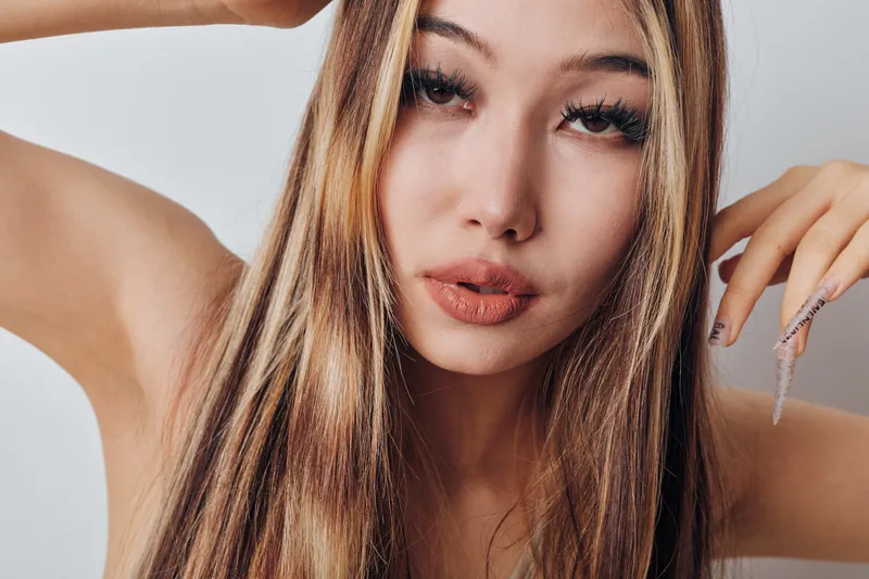 Portrait of young woman with long straight hair posing with raised hands, natural look and stylish makeup on light background in studio. Closeup beauty shot of female face and hair.