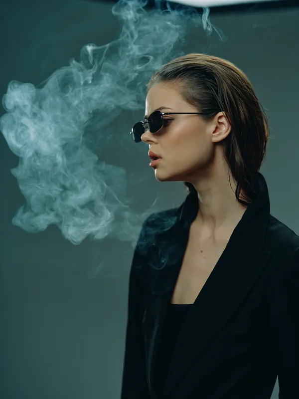 Stylish woman with sunglasses exhaling smoke in dark elegant outfit, moody lighting, and modern atmosphere.