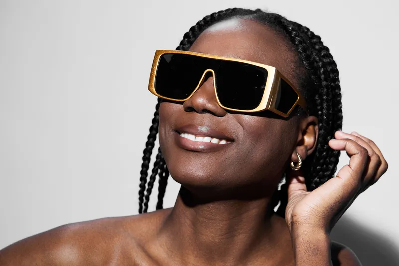 Stylish African woman with braided hair wearing oversized gold sunglasses, smiling confidently against a minimalist gray background, evoking a sense of modern fashion and empowerment