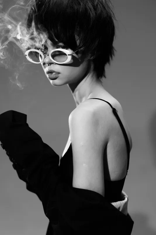 Black and white portrait of a stylish woman wearing sunglasses exhaling smoke with a moody expression in a minimalist setting.