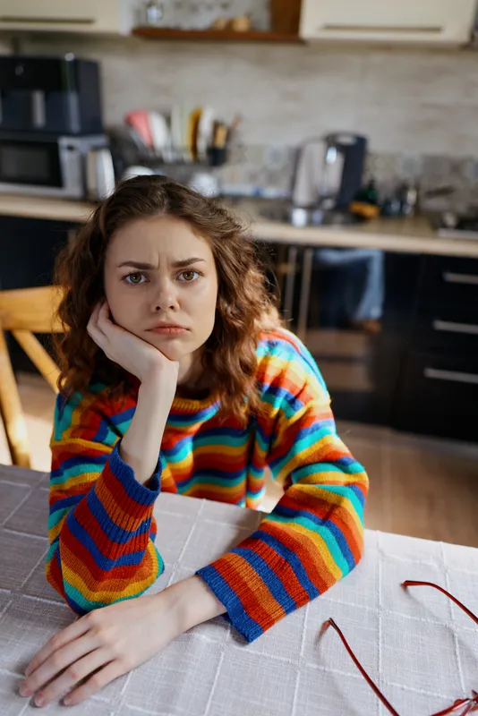 A woman sits at a kitchen table, chin resting on her hand, wearing a colorful striped sweater. A calm, contemplative moment reflecting everyday home life.