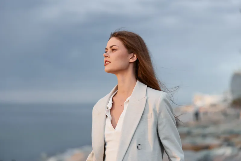 Woman in light blazer standing confident by the sea with wind blowing her hair, natural makeup, and cloudy sky background for fashion theme