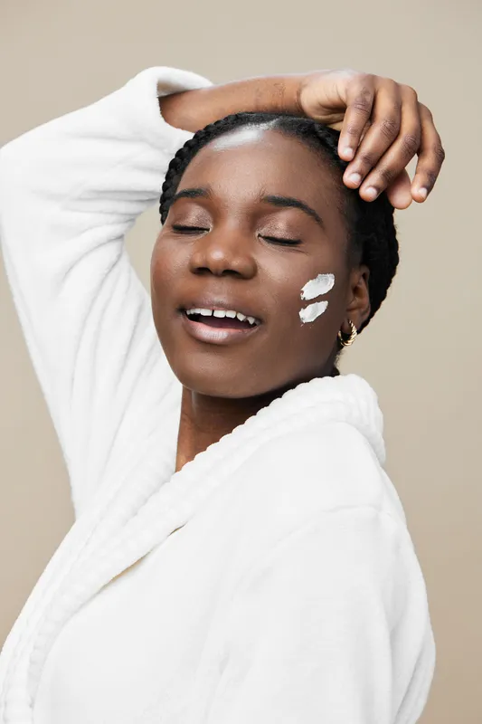 Beautiful young woman enjoying skincare routine, wearing a white robe and smiling, with facial cream on her cheek, against a neutral beige background