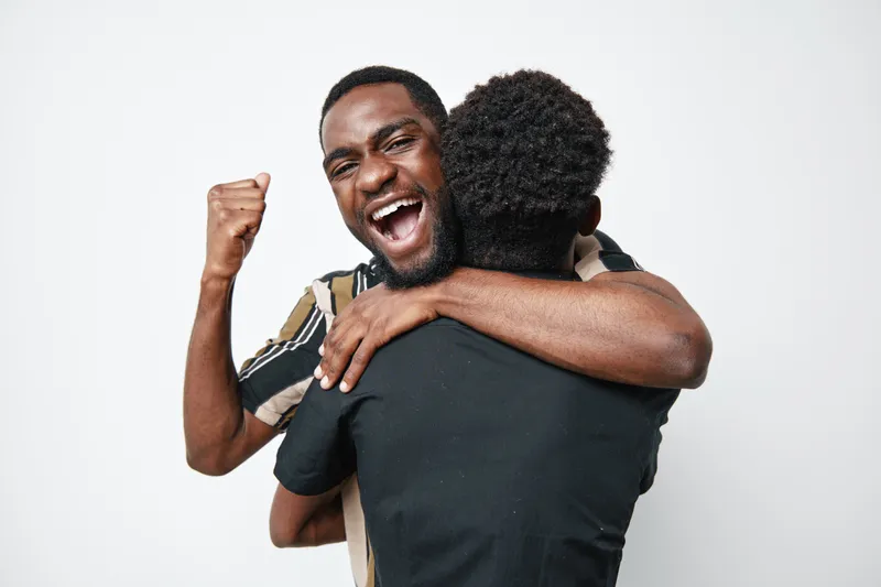 Two black men hugging and celebrating with joy on white background. African ethnicity, dark skin, happiness, friendship, emotional connection, casual clothing, positive energy.
