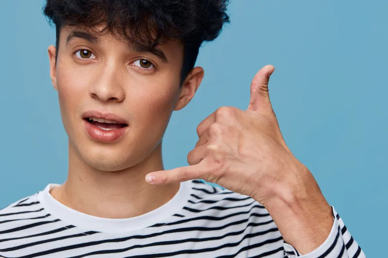 Young man making a call gesture with his hand, wearing a striped shirt, with a surprised expression against a solid blue background.