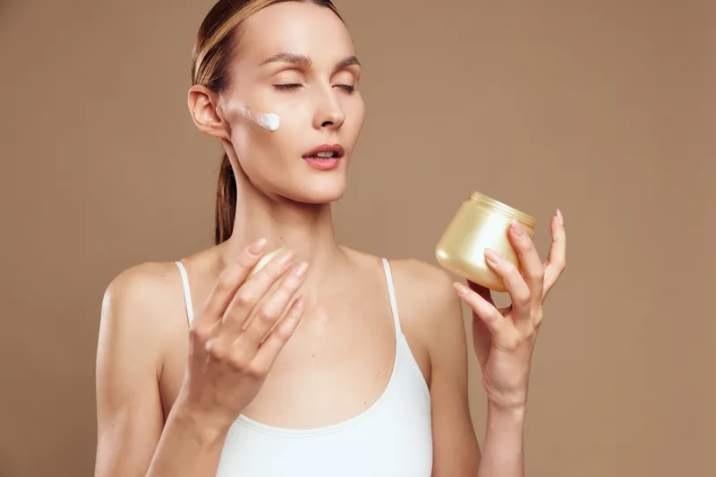 Young woman with closed eyes applying face cream on cheek holding jar in hand. Skincare routine concept with natural makeup and soft lighting on beige background.