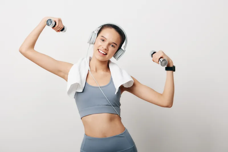 Woman fitness enthusiast exercises with dumbbells, wearing a sports bra and headphones, towel draped, performing lively arm movements in a bright studio for a dynamic workout scene.
