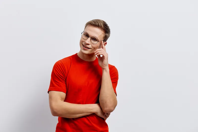 Smiling woman with short brown hair and glasses wearing a bright red t-shirt poses confidently against a plain light gray background. She appears thoughtful and cheerful, creating a friendly and appro