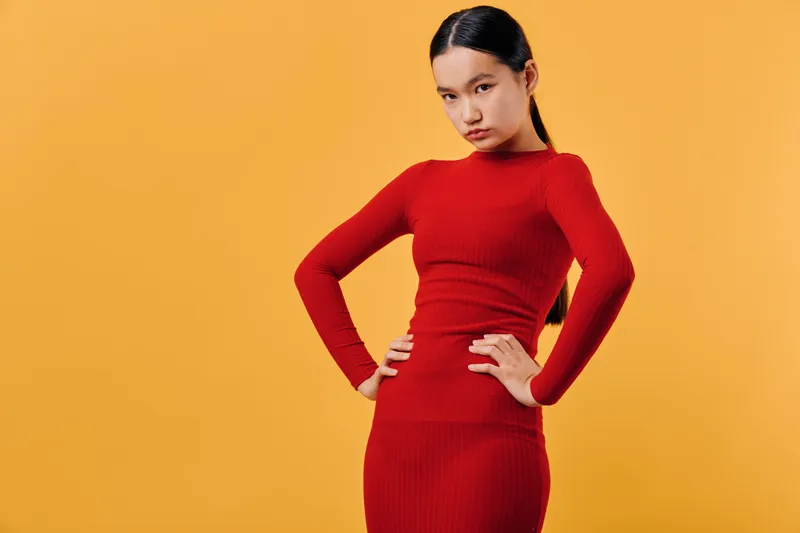 Young woman in red dress posing with confident expression and hands on hips against orange background. Casual style, fashion, portrait and body language with serious mood and modern look.