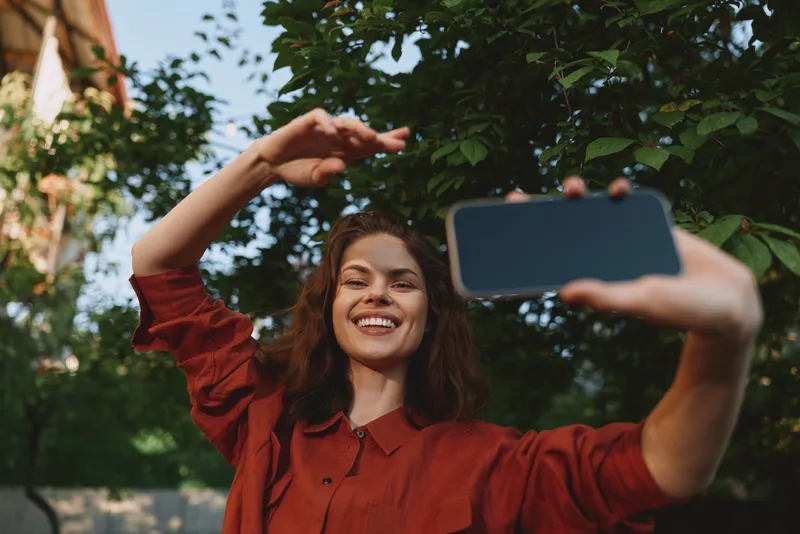 Smiling woman in a red shirt taking a selfie outdoors. Closeup with lush greenery in the background, conveying lifestyle, joy, and self expression in bright daylight