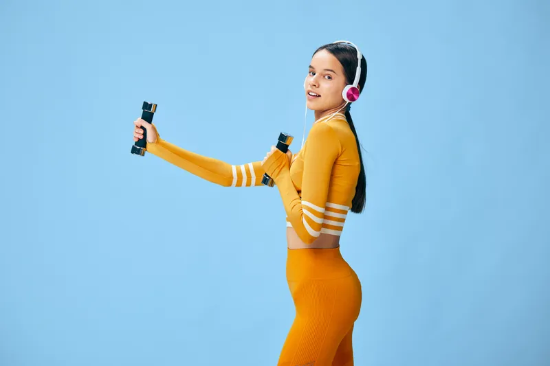Young woman in orange activewear lifts light dumbbells while wearing pink headphones, smiling confidently in a dynamic fitness pose against a clean light blue studio background.