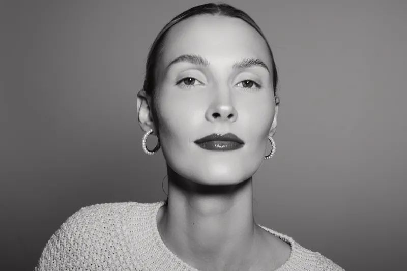 Portrait of a confident white woman with light skin wearing hoop earrings and knitted sweater on grey background.