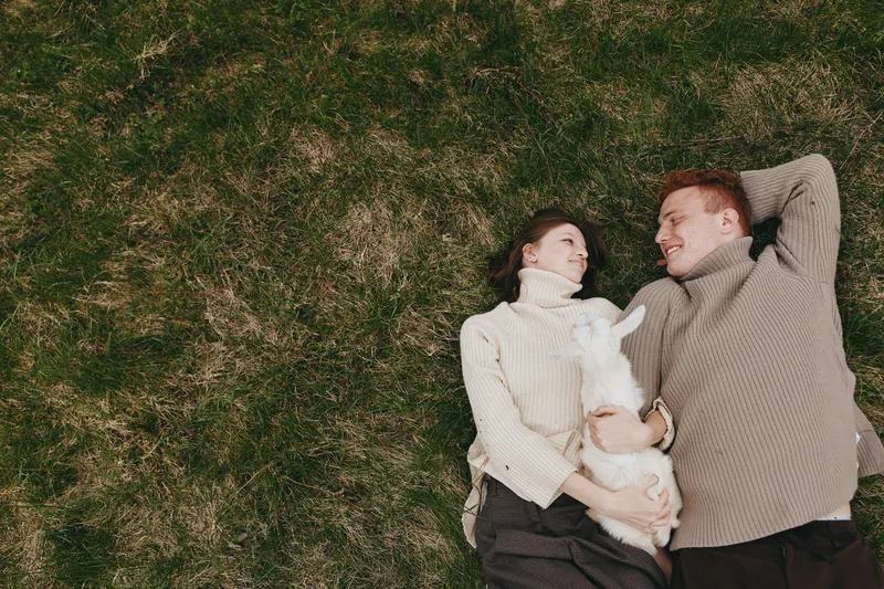 A relaxed couple lies on grass with a playful goat kid nearby, enjoying a carefree outdoor moment. The scene captures simple affection, family bonds, and nature inspired lifestyle.
