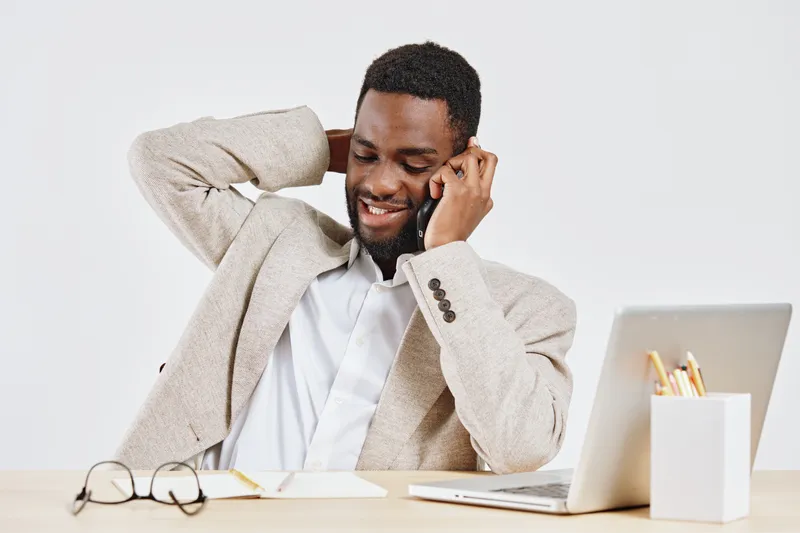 Smiling man wearing beige cardigan and white shirt, talking on mobile phone, sitting at desk with laptop, glasses and notebook, healthy hair, positive mood, modern office setting, lifestyle concept