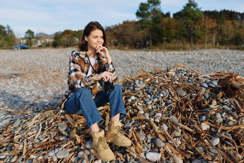 Casual woman sits on a shell strewn beach, deep in thought, wearing a plaid shirt, jeans and boots, against a calm coastal backdrop and open sky.