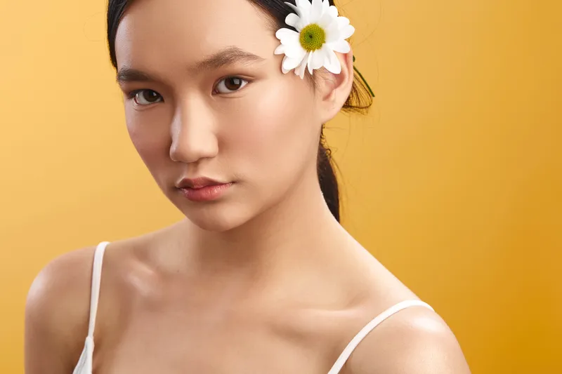 asian woman with a delicate flower in her hair gazes softly at the camera, a clean studio setting emphasizing natural beauty and calm confidence.