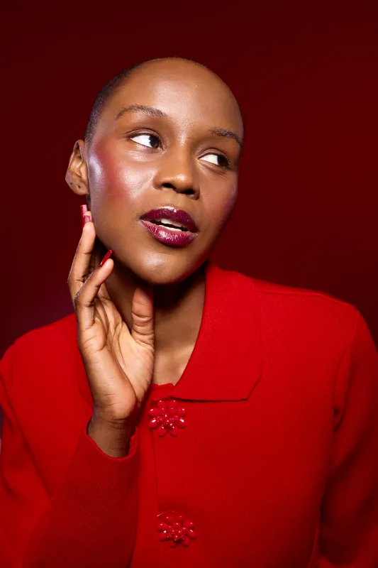Portrait of a dark skinned African woman with shaved head wearing red sweater, looking away thoughtfully. Studio lighting highlights her smooth skin and glossy lips. Beauty concept.