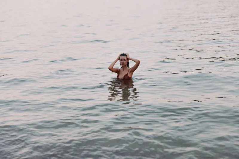 Woman standing in calm water at sunset, wearing a swimsuit, looking thoughtful and relaxed with soft natural light and peaceful atmosphere.