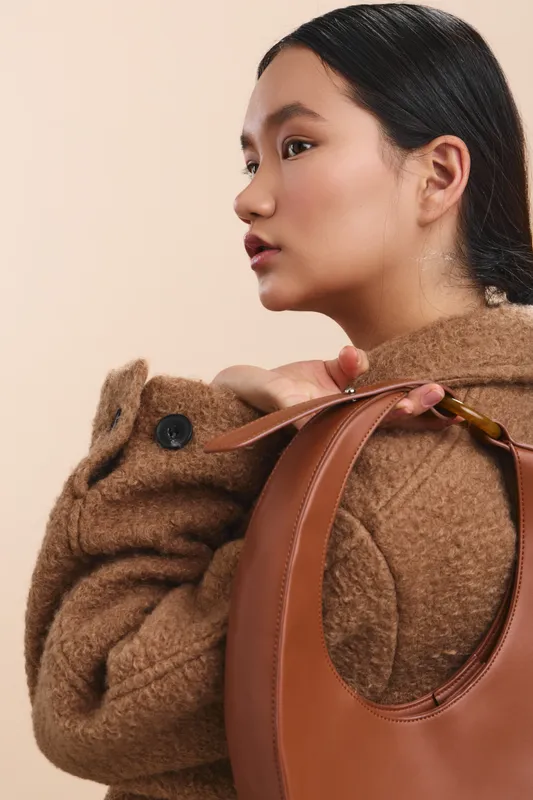 Asian woman wears a warm brown coat and holds a stylish handbag in a neutral studio. She looks away with a calm smile, projecting confident, refined fashion and modern ease.