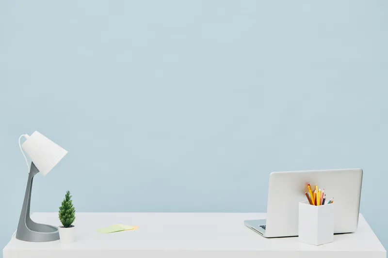 Modern minimalist workspace with a sleek white desk, a laptop, a small potted plant, a white pen holder filled with colorful pens, and a stylish desk lamp on a pastel blue background. Technology and o