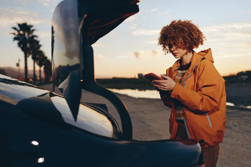 Woman lifestyle outdoors by car at sunset, young curly hair, warm orange jacket, street moment, gentle breeze, relaxed posture, golden sky light, cinematic mood