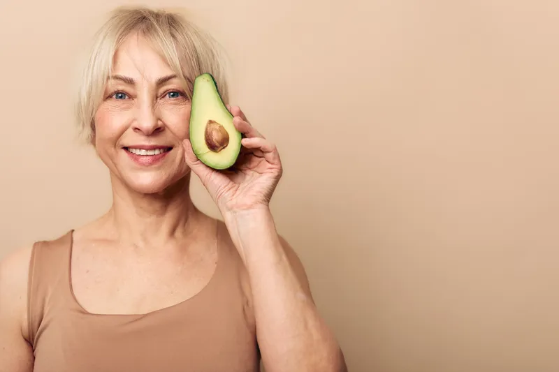 Smiling senior woman holds half of fresh avocado near face on beige background. Concept of healthy lifestyle, nutrition, natural food, and positive aging with clean minimal composition.