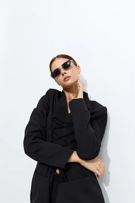 Fashion woman posing with black sunglasses and a stylish black jacket against a minimalist white background, showcasing modern chic and confidence.