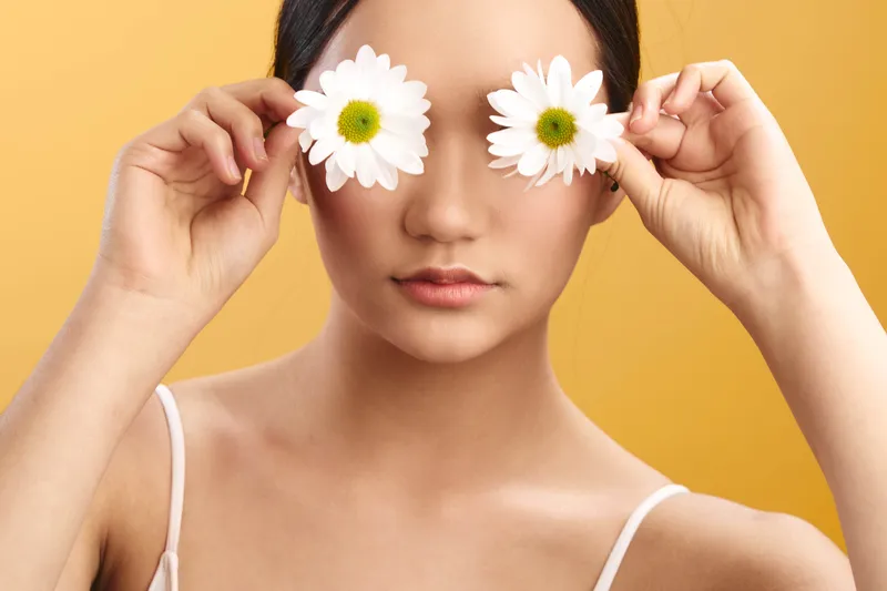 Asian woman holds white daisies over her eyes, creating a playful yet serene portrait. The soft lighting and warm backdrop evoke whimsy, beauty, and calm, inviting viewers to pause and appreciate