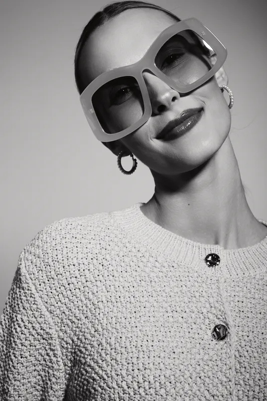 Portrait of a stylish white woman with fair skin wearing oversized sunglasses and hoop earrings. She smiles confidently in a textured sweater against a plain background.
