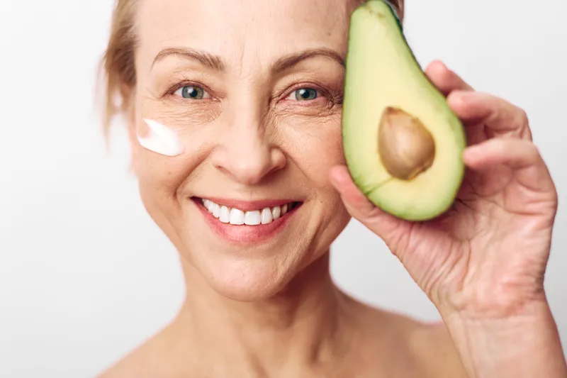 Smiling middle aged woman with natural skin holds fresh avocado and applies white cream on cheek, isolated on white background. Concept for skincare, healthy lifestyle, and natural beauty care.